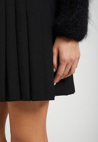 Zwarte geplooide rok met een gestructureerde afwerking. Hand rust zachtjes op de stof, wat de strakke lijnen en de gestructureerde vormgeving benadrukt.