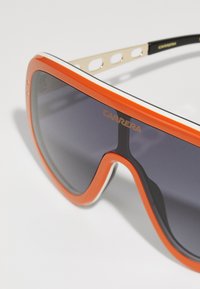 Occhiali da sole over-sized arancioni e neri con lenti scure sfumate. Presentano una montatura con accenti dorati e dettagli bianchi lungo la parte superiore.
