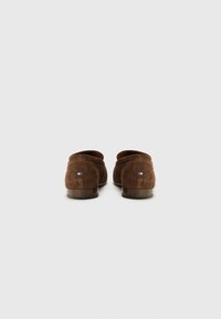 Chaussures en daim marron à enfiler avec un design incurvé, des talons bas et une petite étiquette de logo en rouge, blanc et bleu sur le côté extérieur.
