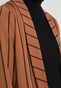 Braun gestreifter Blazer mit schwarzen Linien, der eine glatte Textur und ein offenes Vorderdesign aufweist. Trägt man über einem schwarzen Rollkragenpullover.