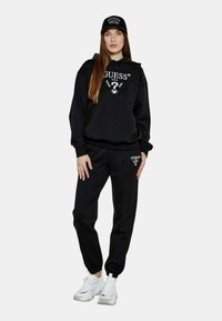 Svart huvtröja och sweatpants-set med vit "GUESS"-logotyp. Slät tyg, avslappnad passform, ribbad mudd. Kombinerat med vita sneakers för träning och en svart keps.
