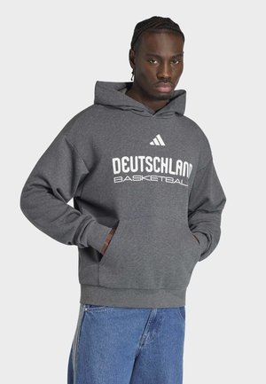 Man met gevlochten haar draagt een grijze hoodie met de tekst "Deutschland Basketball" en het Adidas-logo, gecombineerd met een blauwe spijkerbroek, handen in de zakken.