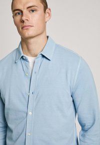 Camisa azul claro de botones con cuello estándar, mangas largas y un tejido suave y texturizado. Botones en un color contrastante.