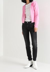 Roze capuchontrui, witte logo t-shirt, zwarte skinny jeans en glanzende witte enkellaarzen met een sleehak.