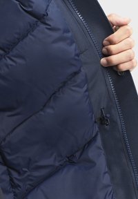 Navy pufferjack interieur met gewatteerde vulling, soepele textuur, zichtbare rits en een trekkoord met een bevestiging voor verstelbare pasvorm.