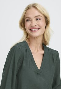 Femeie blondă cu părul lung zâmbește ușor, purtând o bluză verde închis cu decolteu în V, pe un fundal deschis simplu.