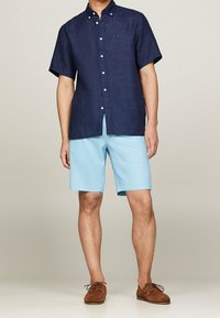 Navy short-sleeve button-up shirt paired with light blue shorts. Model wears brown moccasin shoes, standing against a neutral background.