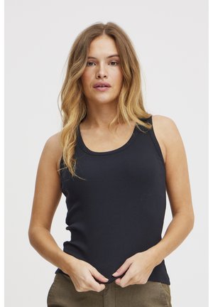 Frau mit langen blonden Haaren, die ein schwarzes ärmelloses Oberteil trägt und am Saum gegen einen einfarbigen weißen Hintergrund zieht.