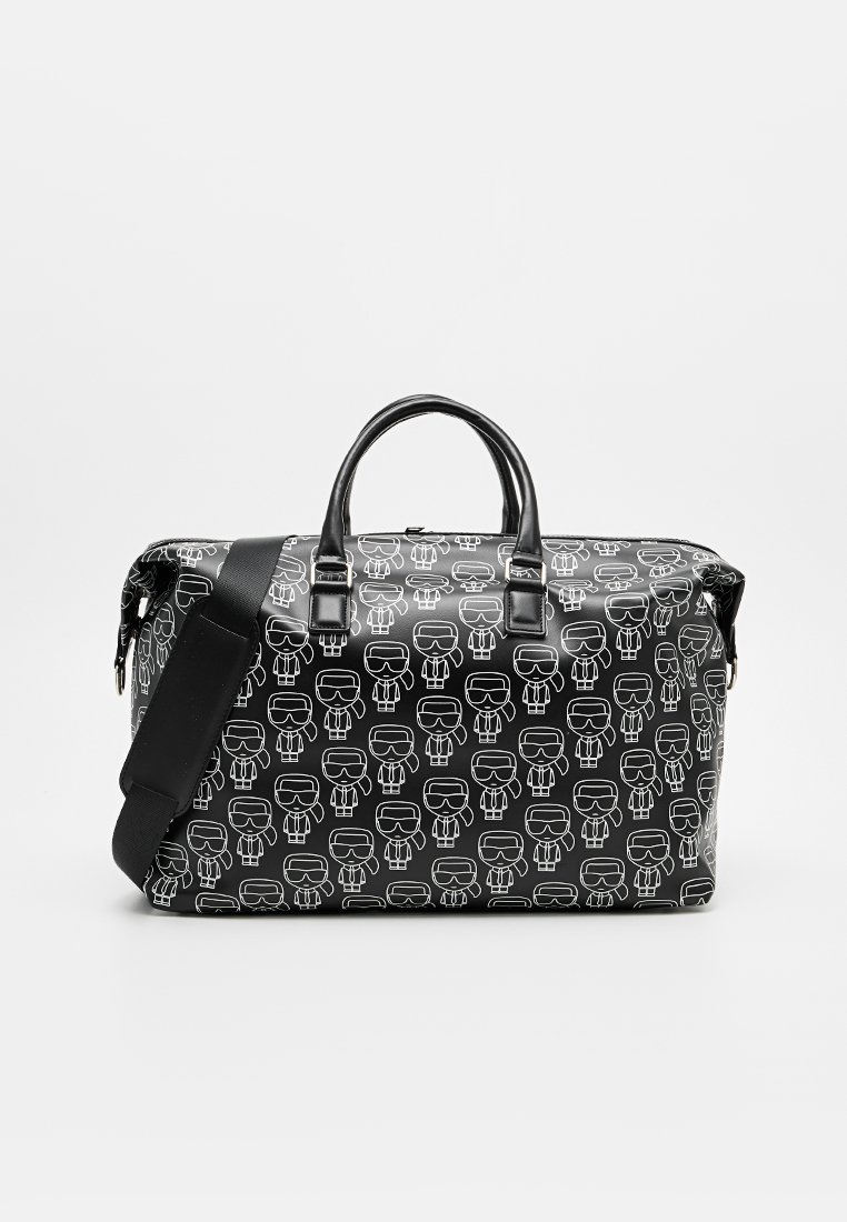 Bolsa de lona negra con patrón de figuras de dibujos animados blancas, cuenta con dos asas negras, correa de hombro desmontable y cierre de cremallera.