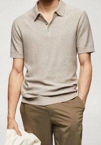 Hombre vistiendo una camiseta de polo de manga corta texturizada beige, ligeramente metida en unos pantalones marrones, sosteniendo una prenda blanca con la mano izquierda.