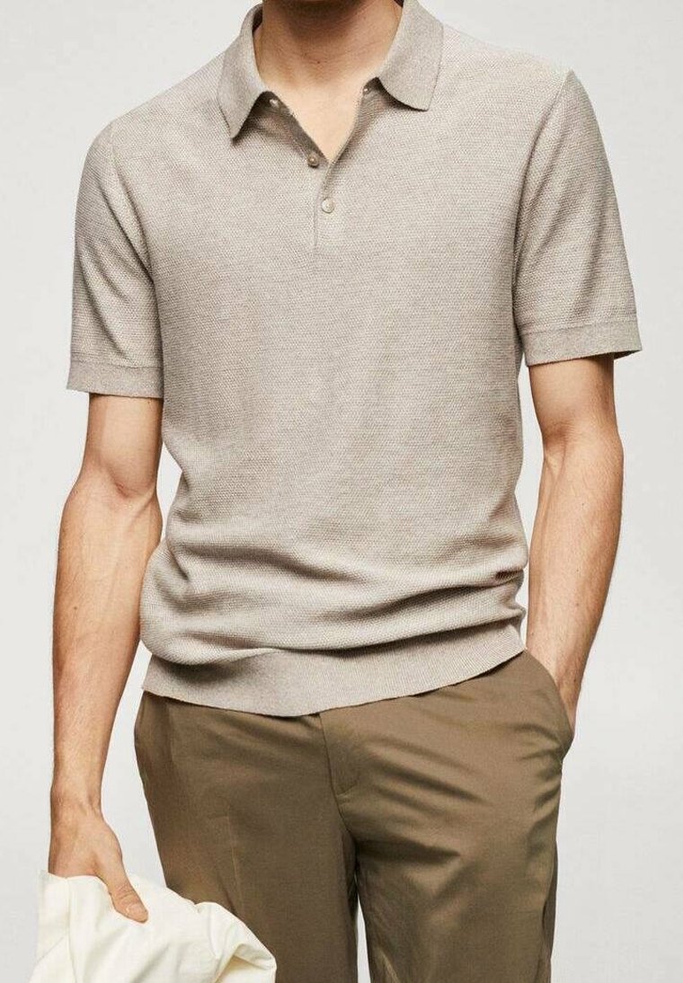 Hombre vistiendo una camiseta de polo de manga corta texturizada beige, ligeramente metida en unos pantalones marrones, sosteniendo una prenda blanca con la mano izquierda.