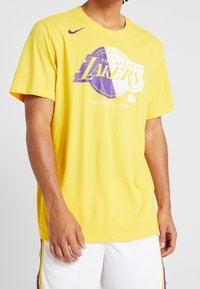 Żółta bawełniana koszulka z fioletowym tekstem "Los Angeles Lakers" i motywem koszykówki. Krótkie rękawy i okrągły dekolt.