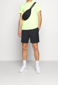 Neon žluté tričko, černé šortky a černá křížová taška. Model má na sobě bílé tenisky a sportovní ponožky, postavený proti neutrálnímu pozadí.