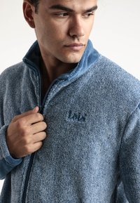 Chaqueta de forro polar en tonos azules, con un alto cuello, cierre con cremallera y tejido texturizado. Presenta un sutil logo grabado en el pecho.