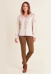Pullover crema con ricamo geometrico rosa, scollatura a V, maniche a palloncino, abbinato a pantaloni skinny marroni e stivaletti. Tessuto liscio.