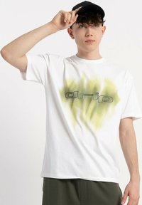 Młody mężczyzna w białej koszulce z zielono-żółtym nadrukiem „drip”, w ciemnych spodniach i czapce w kolorze czarnym, poprawiający czapkę prawą ręką.