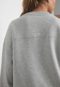 Hallhalline jakk featuring pehmea kude, ümmargune kaelus ja eristuv "self." tikand detail taga.