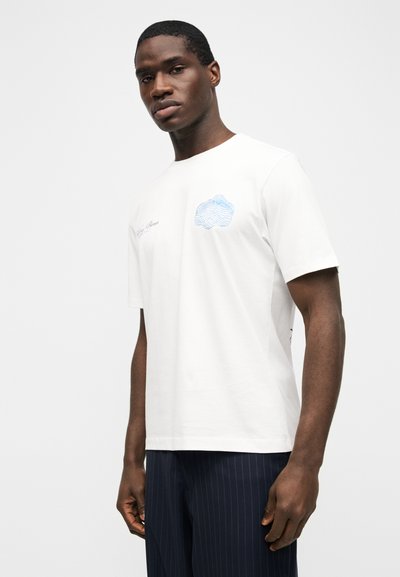 Άνδρας που φοράει λευκό κοντομάνικο T-shirt με μπλε γραφικό στο στήθος και σκούρο μπλε παντελόνι με ρίγες, στέκεται μπροστά σε απλό ανοιχτό φόντο.