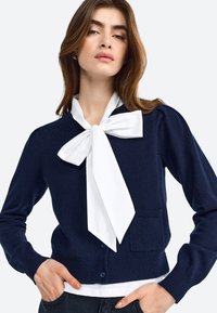 Marinefarbener Cardigan aus Strickstoff mit einer großen weißen Schleife am Kragen, gerippten Bündchen und einer Detailtasche auf der Vorderseite.