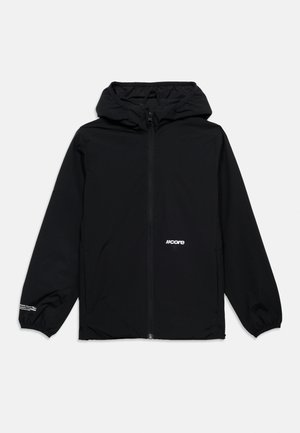 Lächelnder rothaariger Junge, der eine schwarze Kapuzenjacke, blaue Jeans und schwarze Sneakers trägt und vor weißem Hintergrund steht.