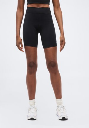 Femeie purtând pantaloni scurți negri de ciclism cu talie înaltă, șosete albe și adidași albi, stând împotriva unui fundal simplu.