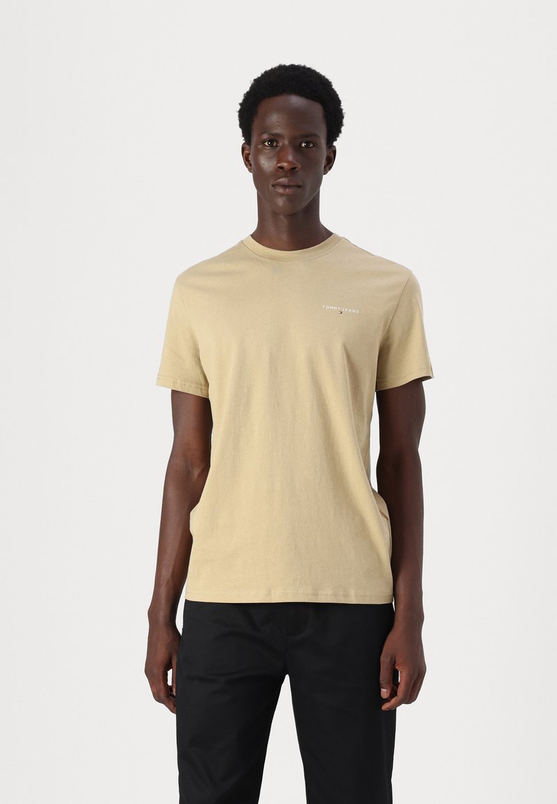 Light beige cotton t-shirt with a crew neck, short sleeves, and a small logo on the chest, paired with black trousers.