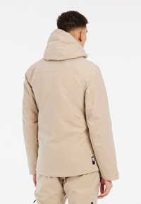 Beige waterdichte jas met capuchon, voorzien van een gladde textuur, ritsluiting en logo-patch op de onderzijde links.