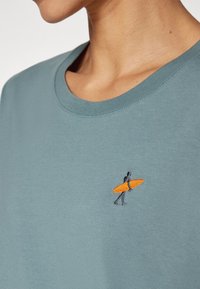 Gray-blue t-shirt with a crew neck, featuring a small embroidered design of a person with an orange surfboard on the chest.