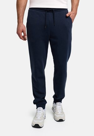 Bărbat purtând pantaloni sport bleumarin cu șnur în talie și adidași albi, stă în picioare cu o mână în buzunar.