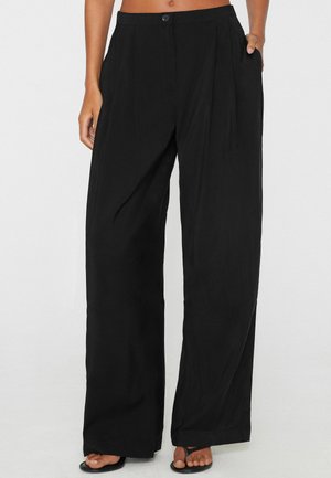 Donna che indossa pantaloni neri a vita alta e gamba larga con tasche e sandali neri con punta aperta su uno sfondo semplice.