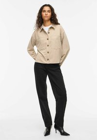 Bēša pogājama jaka ar divām krūšu kabatām, izgatavota no mīksta auduma. Kombinēta ar augsta vidukļa melnām biksēm un ielu sānos spicām kurpēm.