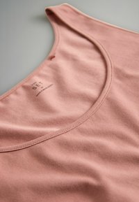 Jasnoróżowy top na ramiączkach z zaokrąglonym dekoltem, gładkiej bawełnianej tkaniny i minimalistycznymi przeszyciami. Wewnątrz przy dekolcie widoczna metka.