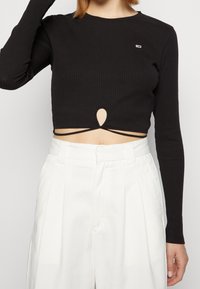Donna che indossa un crop top nero a maniche lunghe con scollatura a chiave e dettaglio incrociato, abbinato a pantaloni bianchi a vita alta con pieghe.