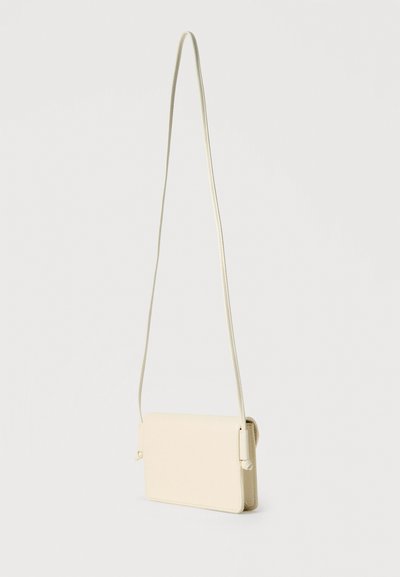Μικρή τσάντα χιαστί από κρεμ δέρμα με μακρύ λεπτό λουρί και ορθογώνιο σχήμα σε απλό λευκό φόντο.