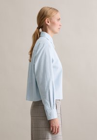 Hele sinine lühendatud nööpidega särk terava kraega ja pikkade varrukatega, mis on kombineeritud ruudulise seelikuga. Sile kangapind.