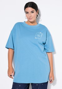Svetle modré oversized tričko vyrobené z měkké látky s kulatým výstřihem. Má malou bílou grafiku na levé části hrudi a volné rukávy.