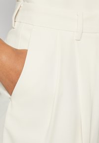 Pantalons ajustés couleur crème avec une texture lisse, des poches latérales et une taille soignée dotée de passants de ceinture.