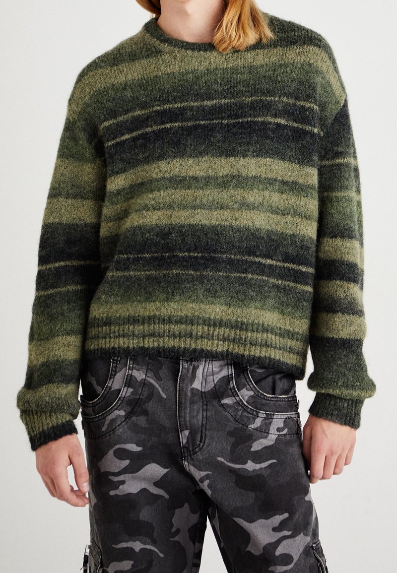 Un pull en tricot rayé vert et gris avec un col rond, des poignets côtelés et une finition texturée, associé à un pantalon cargo camouflage gris.