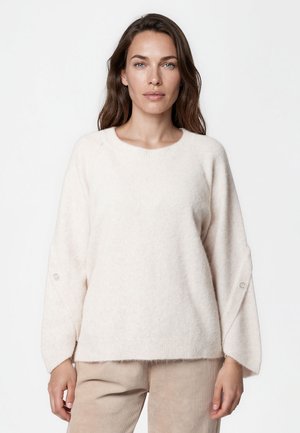 Femme aux longs cheveux bruns portant un pull blanc texturé et ample ainsi qu'un pantalon beige clair, debout devant un fond blanc uni.