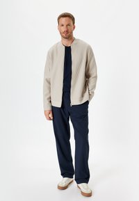 Beige ribbet zip-jakke over en marineblå skjorte, parret med løse marineblå bukser og hvide sneakers med brune accenter. Enkel, moderne design.