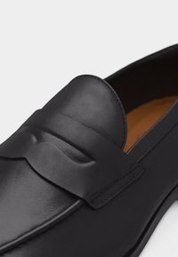 Mocassin en cuir noir avec bout mocassin, bride décorative sur le dessus et doublure intérieure beige.