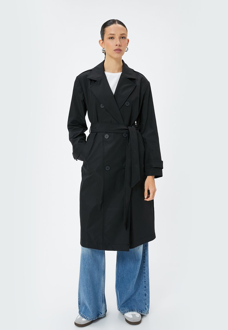 Abrigo negro de trench con botones de doble botonadura y cinturon en la cintura. Combinado con vaqueros de pierna ancha en azul y zapatillas grises. Textura suave.