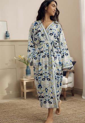 Frau steht barfuß in einem blau-weißen, mit Blumenmuster bedruckten Bademantel, der an der Taille gebunden ist, in einem sanft beleuchteten Wohnzimmer.