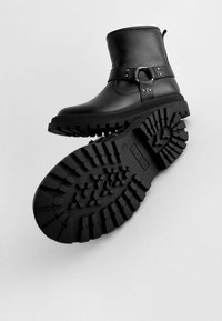 Schwarze Leder-Ankle-Boots mit einer strukturierten Gummisohle. Sie verfügen über einen Riemen mit metallischen Beschlägen, eine runde Zehenpartie und einen niedrigen Absatz.