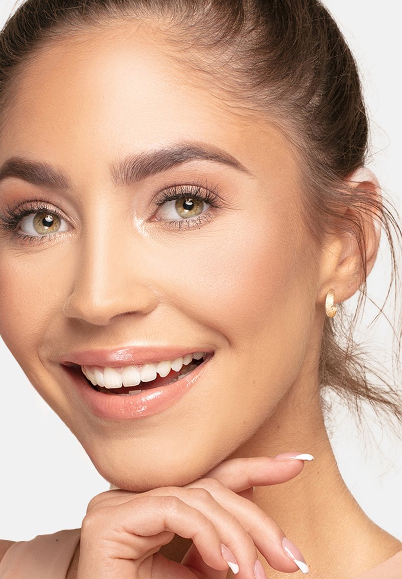 En närbild av en leende person med olivgröna ögon, ljust brunt hår, iförda små guldiga hoops och en mjuk, naturlig sminklook.