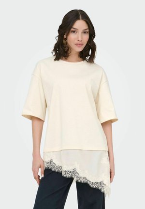 Femme portant un top crème oversize à manches courtes avec un détail en dentelle à l'ourlet et un pantalon noir, debout sur un fond clair uni.