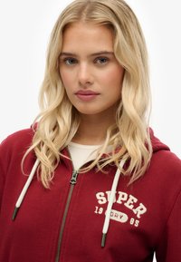 Sudadera con cremallera roja hecha de un tejido suave, con cordones blancos. Presenta el logo "SUPER 1985" en la parte del pecho en color crema. Corte casual, cremallera frontal.