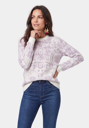 Kvinde iført en hvid og lyse lilla mønstret sweater og blå jeans, med den ene hånd på hoften og den anden, der rører ved hendes hage.