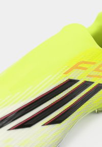 Φωτεινό κίτρινο αθλητικό παπούτσι με υφασμένα επάνω μέρη, γωνιακές γκρίζες και μαύρες ρίγες και κόκκινες λεπτομέρειες, σχεδιασμένο για απόδοση και ευελιξία.