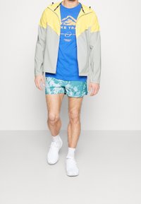 Muž oblečený v žluté a šedé bundě, modrém tričku s potiskem, tyrkysových šortkách s vzorem, bílých ponožkách a bílých teniskách kráčí vpřed.